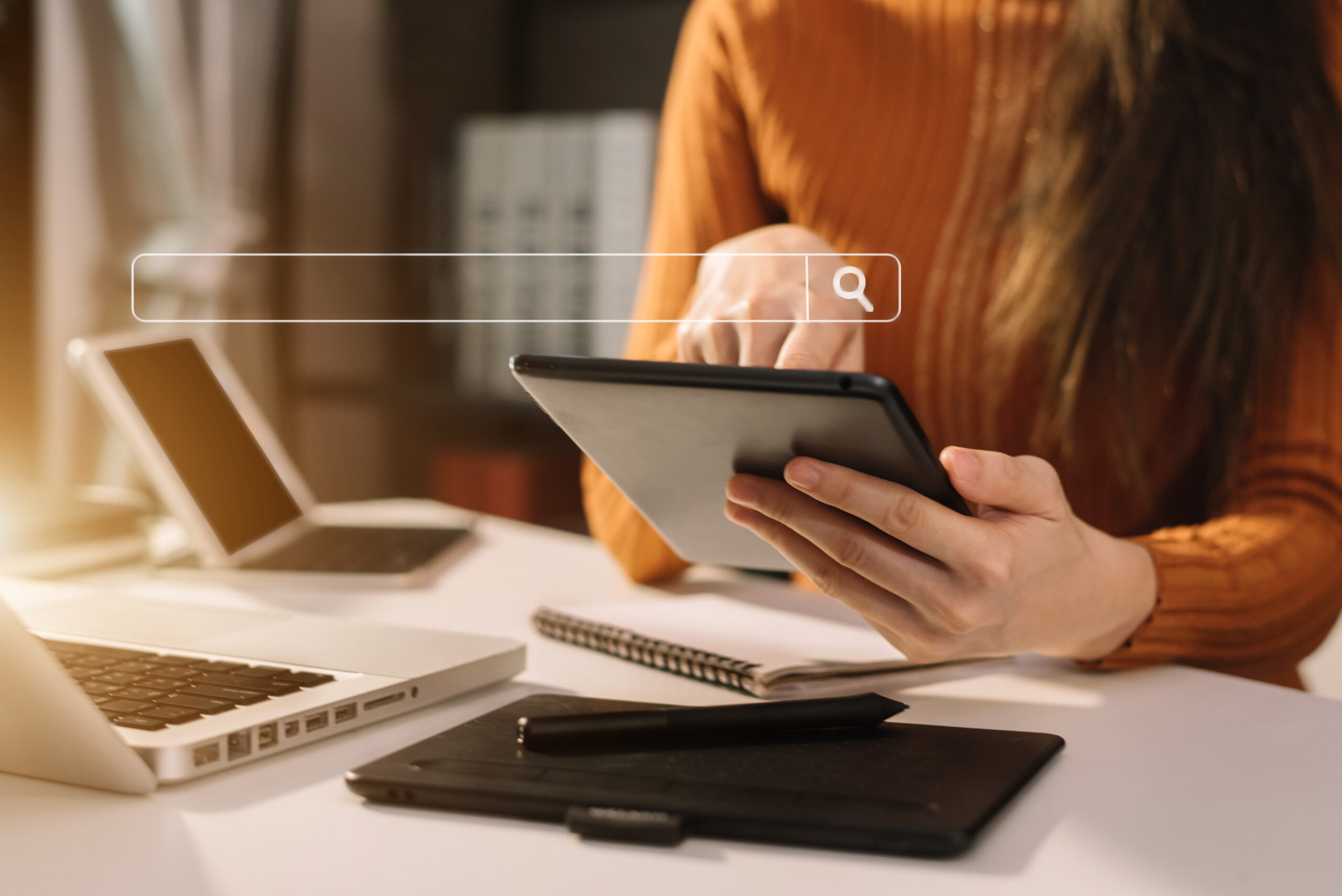 Person using a tablet device at a desk with a search bar graphic overlay. The workspace includes a laptop, notepad, and stylus, suggesting a focus on technology, research, or online search activities.